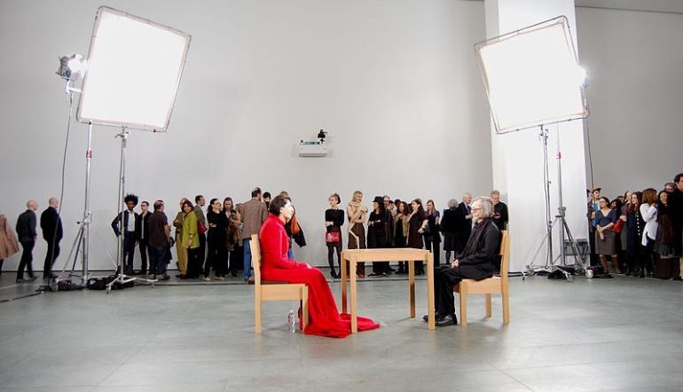 Marina Abramovic The Artist Is Present, Marina in red sits in one chair, a guest sits in a chair across the table