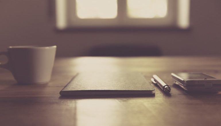 monochromatic image, a desk in front of a lit window with a cup, a notebook, a pen, and a smartphone