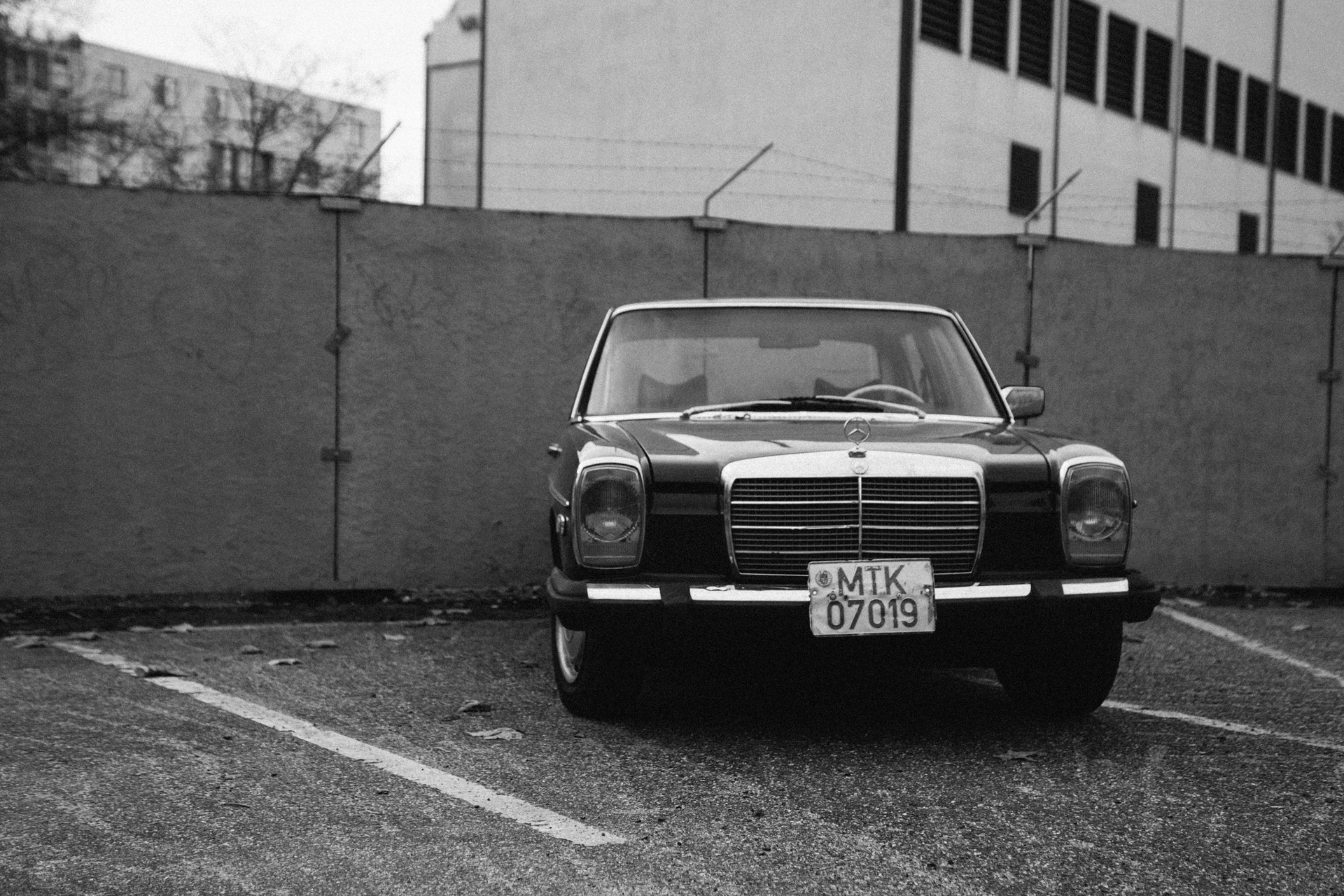 Black and white photograph of an old car in front of a cement wall with barbed wire