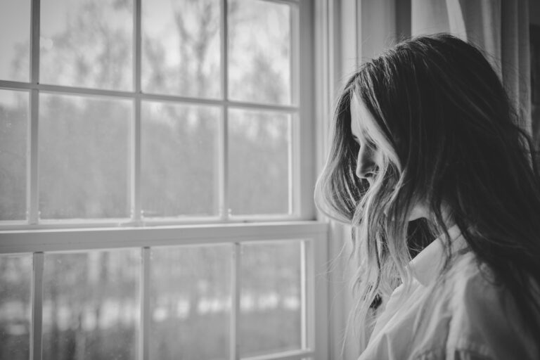 In a greyscale photo, a woman stands in front of a window.