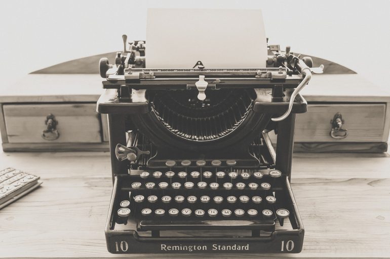 image of a typewriter sitting on a wooden desk - the typewriter is a "Remington Standard" and a blank piece of paper is loaded
