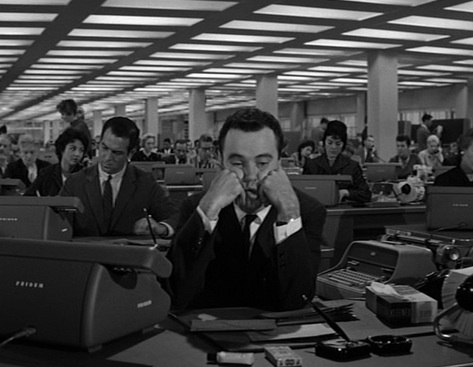 photo of a man sitting a desk in a large office filled with people--he looks exasperated and has his face in his hands