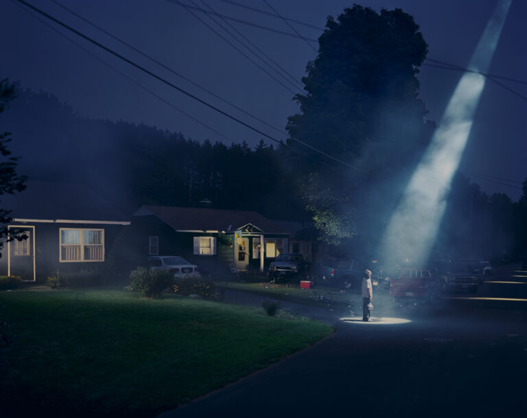 a photograph by the contemporary US photographer Gregory Crewdson of a nighttime suburban scene in which a single figure stands at the end of a driveway with a large beam from some unknown source beaming down on the figure