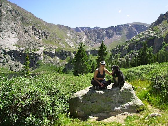 woman and her dog sitting on a rock in the midst of some greenery, trees, and mountains