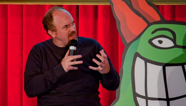 Comedian Louis CK on stage and looking to his left with his hands up
