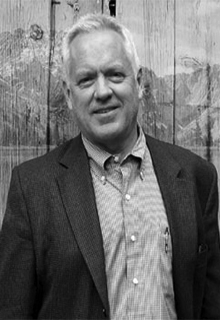 A black and white image of a white man smiling in front of a reflection of mountains