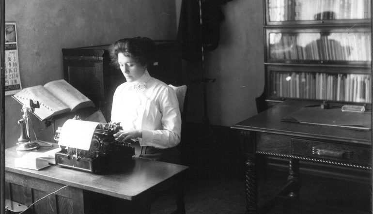 1912 photo of secretary typing at typewriter in front of bookcase