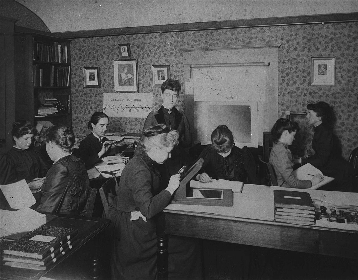 Black and white picture of women in period clothing doing different activities on a sitting room. 