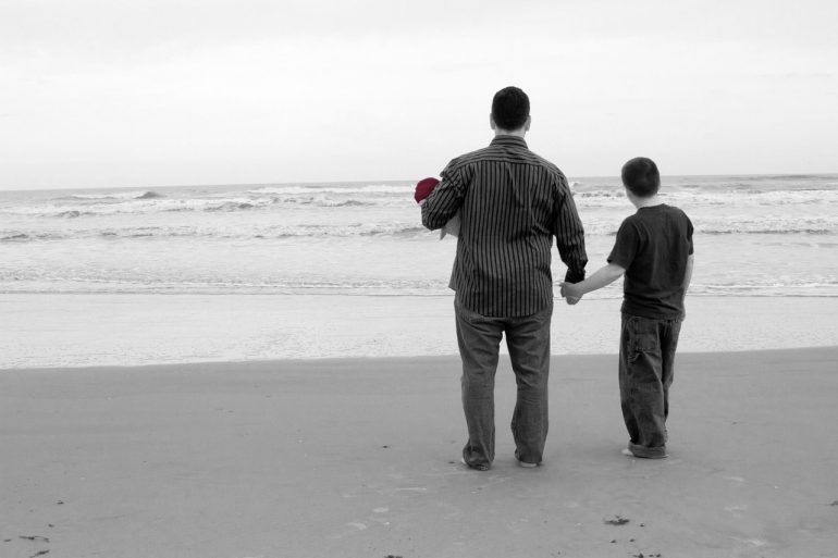 Black and white picture of a man and a boy holding hands on a beach looking away from the camera facing the sea. 
