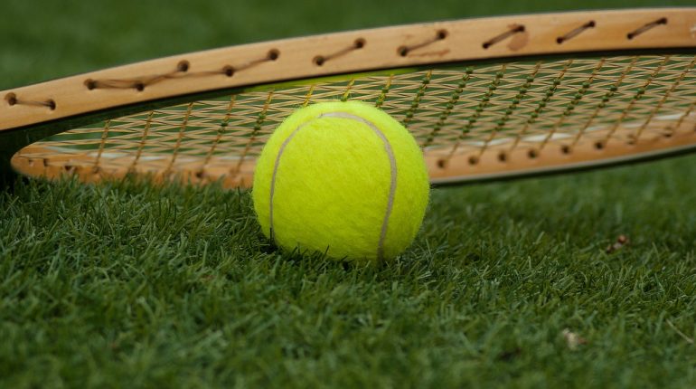 Tennis ball on the grass with a tennis racket lying on top of it.