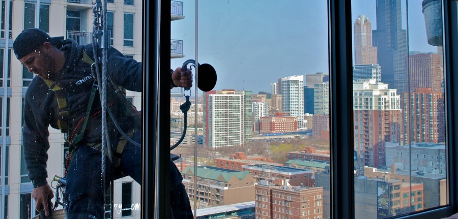 A picture of a window washer taken through the window. In the background you can see a city landscape.  