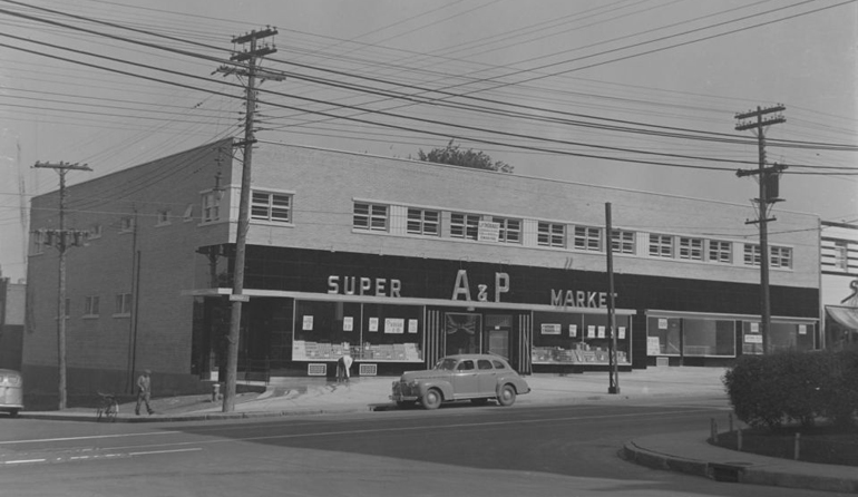 Black and white photograph of an old car parked outside a supermarket