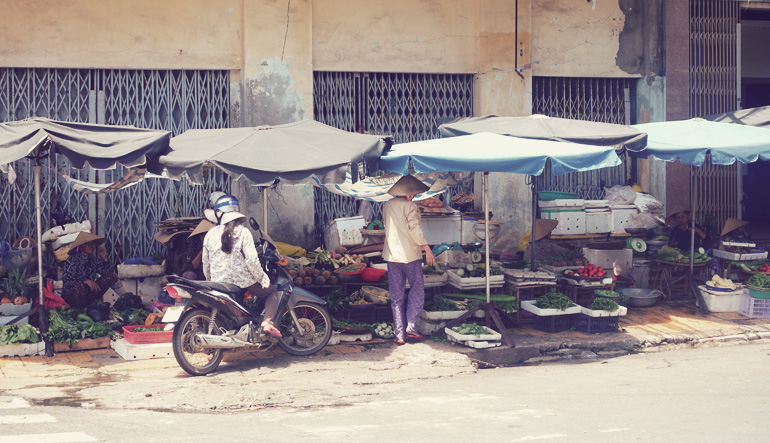 Market stalls for produce with a person on a motorcycle looking at the goods.