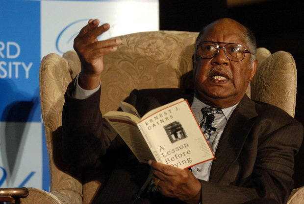 Man with glasses sitting in an armchair reading from "A Lesson Before Dying" by Ernest J. Gaines.