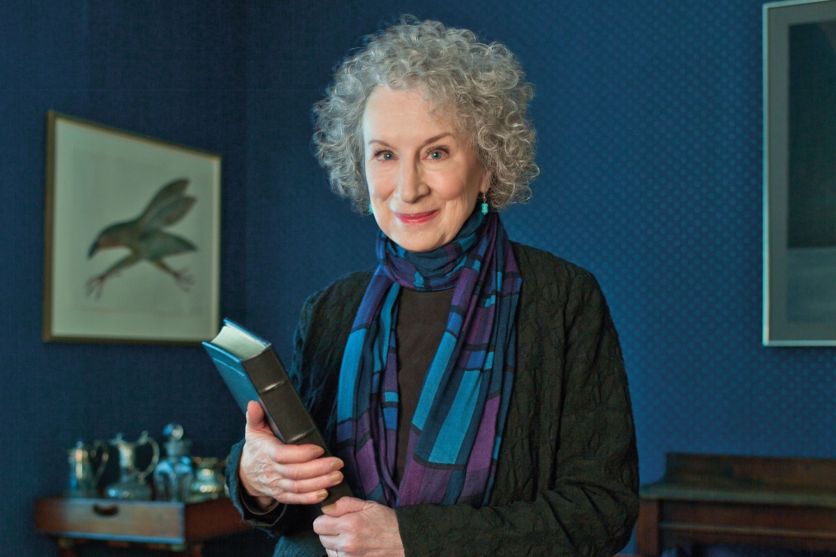 Woman with curly gray hair holding a book and posing for the camera in front of a blue wall.