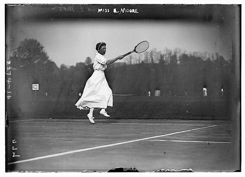 Old photograph of a woman wearing a white dress and holding a racquet.