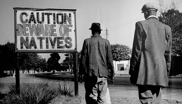 Two men in suits walking towards a sign that reads "Caution Beware of Natives."
