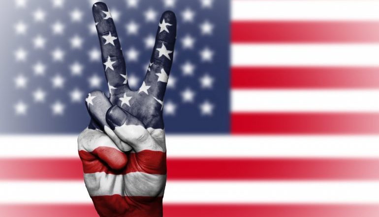 A hand holding a peace sign in front of the American flag.