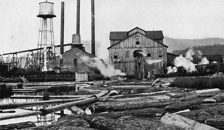 Old photo of a factory with smoke surrounding it, and logs laying on the ground.