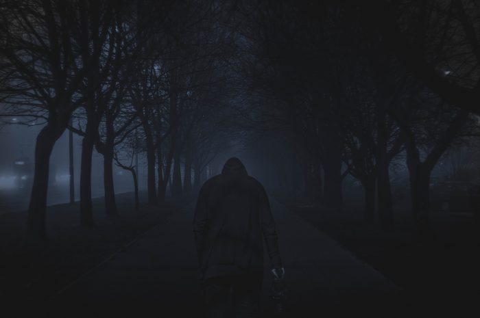 Hooded figure standing in front of a dark walkway lined by trees.