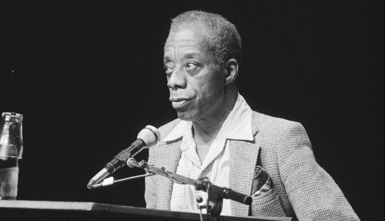 Black and white photo of James Baldwin standing behind a podium speaking into a microphone