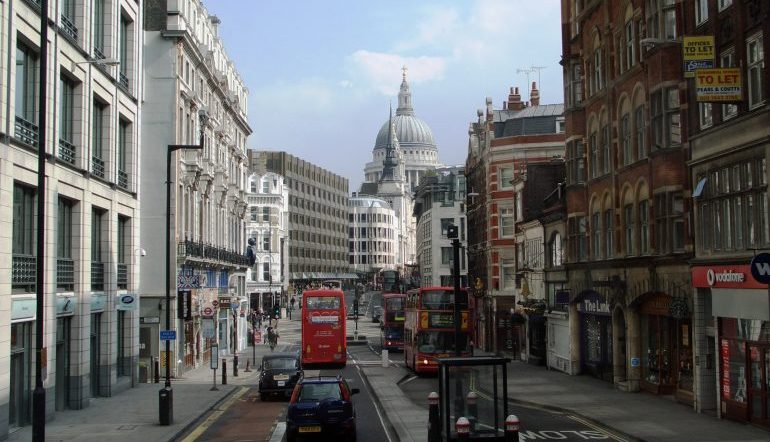 Street in London lined with both older and more modern buildings. A red doubledecker bus on one side and a large domed building in the skyline.