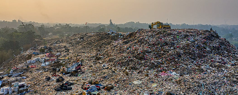 a bird's-eye photograph of a massive pile of trash, to the right hand side construction equipment sits idle; the beyond the pile is a foggy jungle