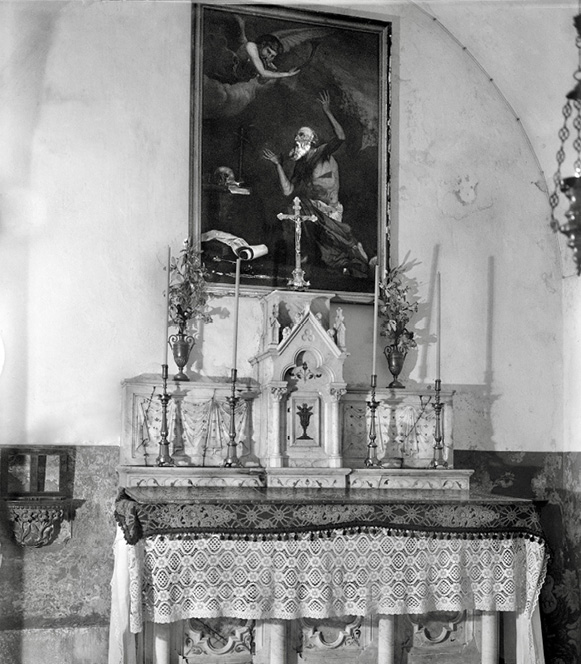 A black and white photograph of an altar with a large oil painting behind it of a frenzied, half-naked man next to a human skull set on a writing table.