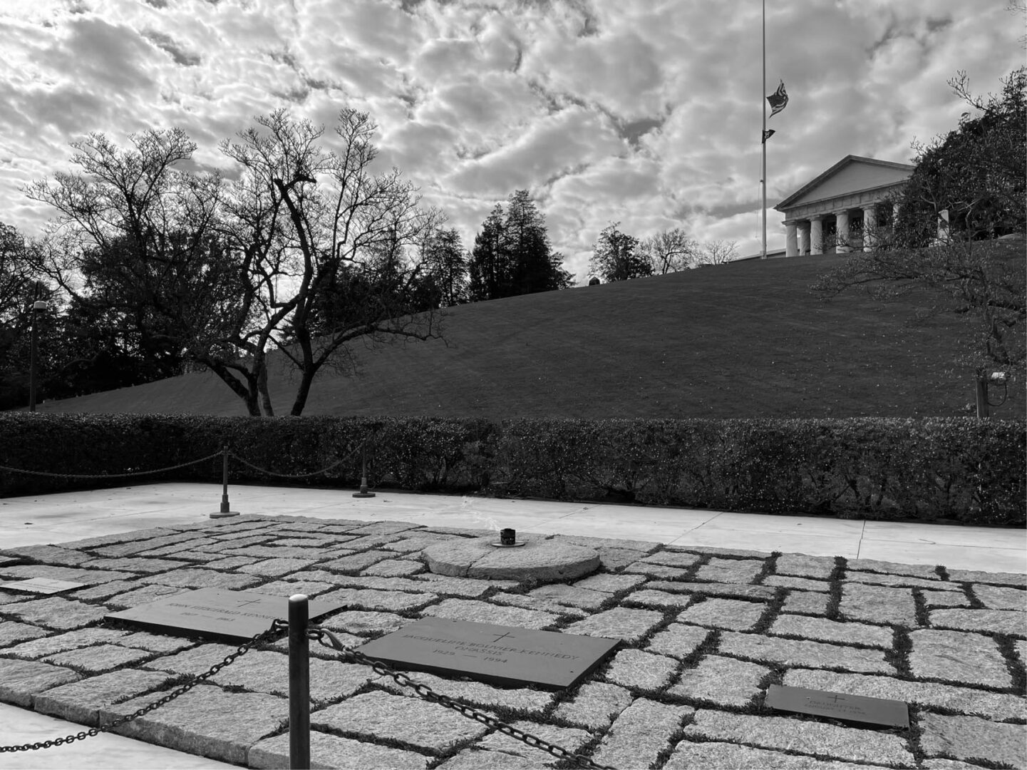 Black and white photo of a cemetery with stone slab grave markers, a neoclassical building in the background, and the American flag at half-mast.