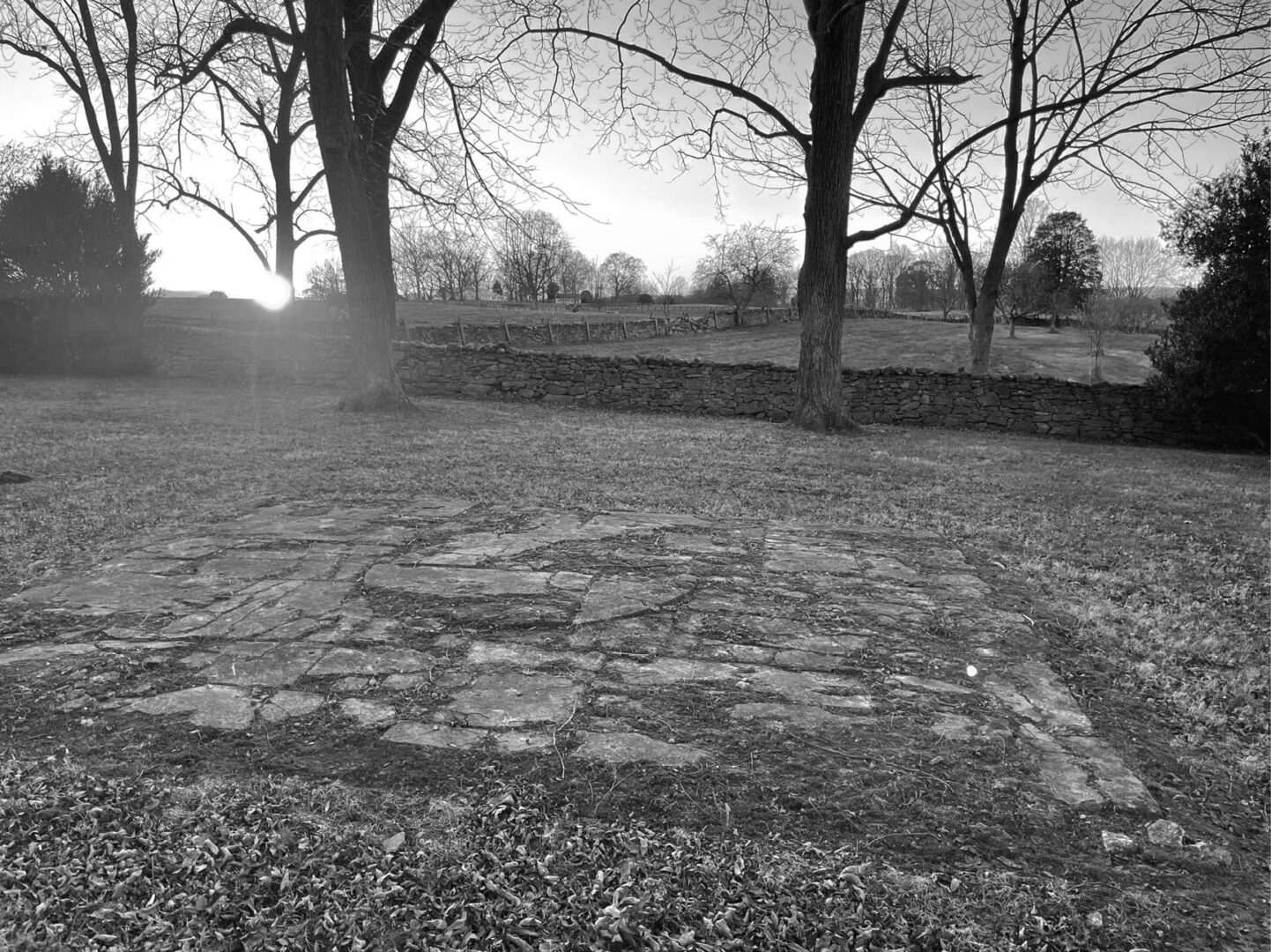 Black and white photo of a stone slab on grass, with trees and a stone wall in the background during sunset or sunrise.