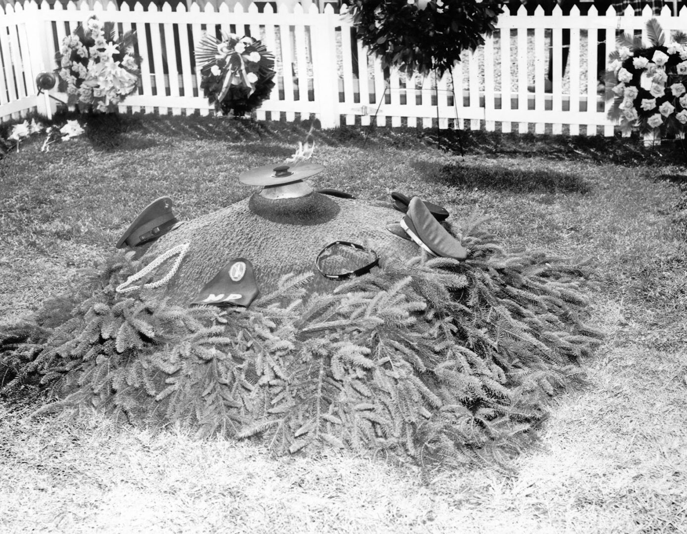 A grave decorated with branches and military hats with a white picket fence and wreaths in the background.