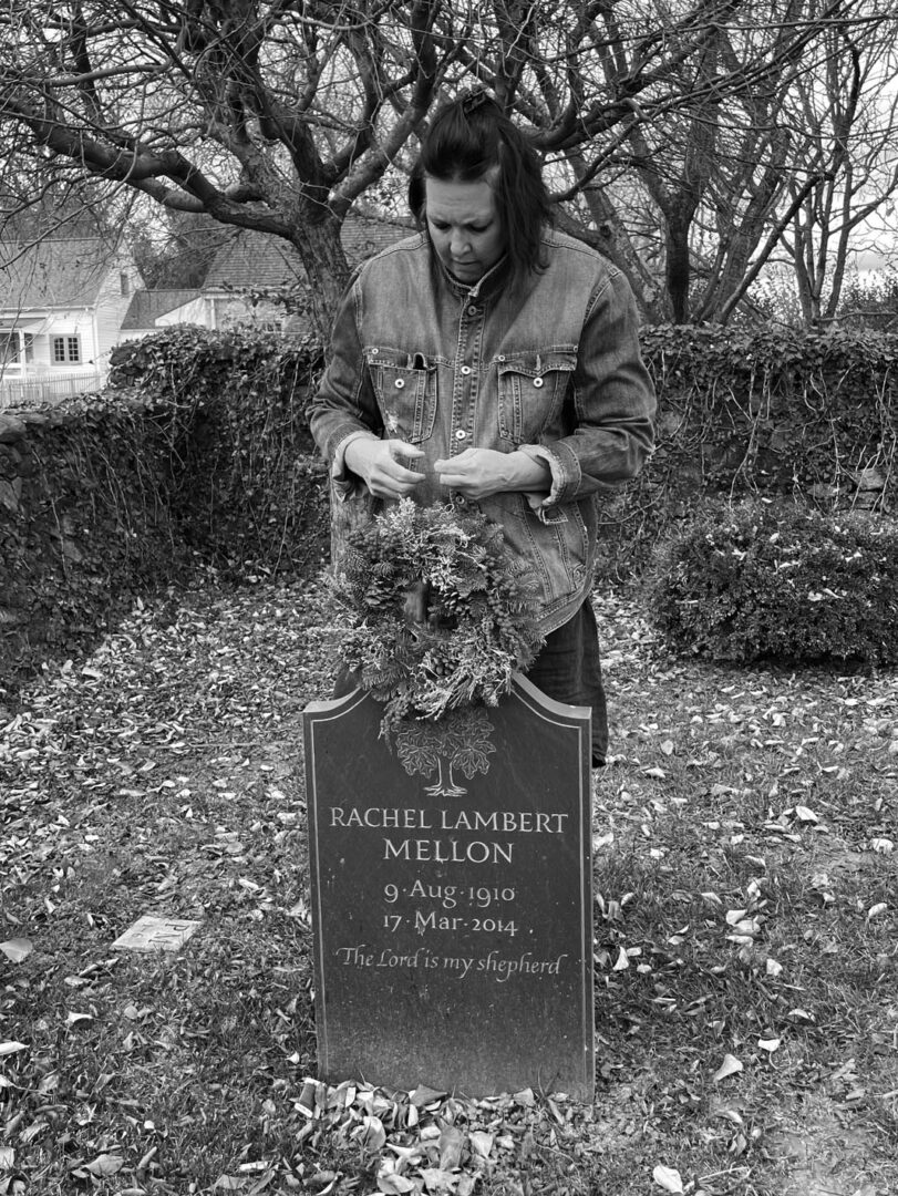 Photo of a woman behind a grave stone, placing a wreath on top. The grave says "Rachel Lambert Mellon, 9 Aug 1910 - 17 Mar 2014, the Lord is my shepherd."